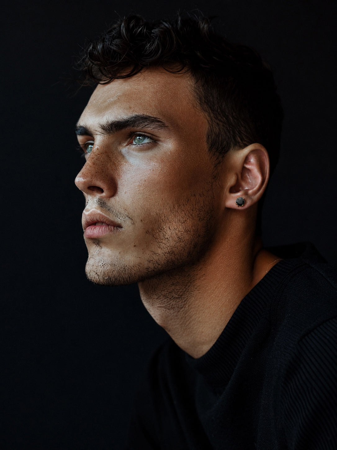 Ein junger Mann mit kurzen dunklen Haaren und hellen Augen blickt vor einem dunklen Hintergrund nach links; er trägt einen schwarzen Pullover und präsentiert auffälligen Bruttall-Herrenschmuck, der mit Swarovski-Zirkonia verziert ist.