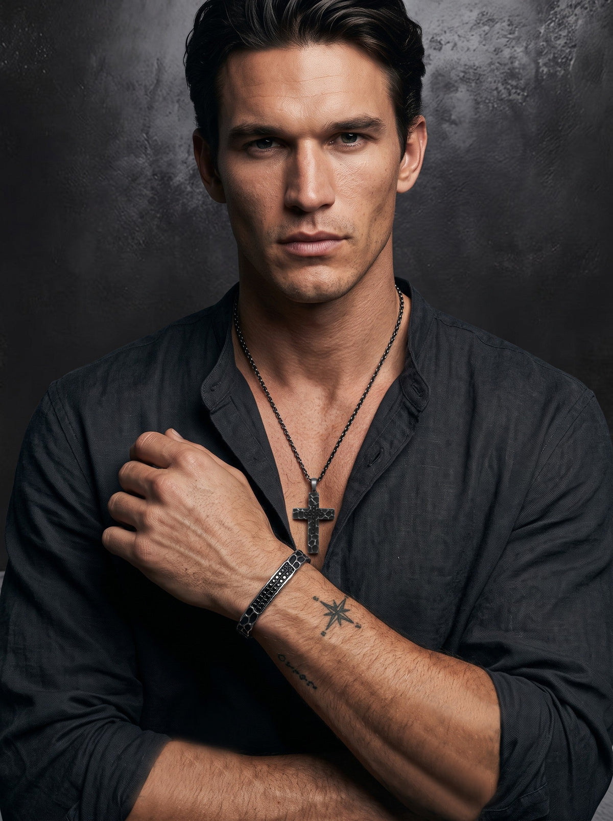 A man wearing a dark shirt poses against a textured dark background, showing a cross necklace, a bracelet, and a star tattoo on his forearm.