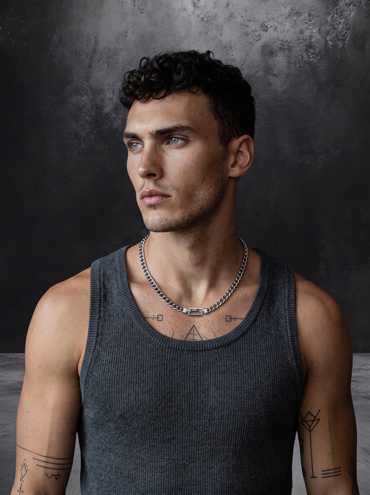 A man with short curly hair, wearing a gray sleeveless shirt, silver chain necklace, and visible arm tattoos, stands against a dark textured background, looking to the left.