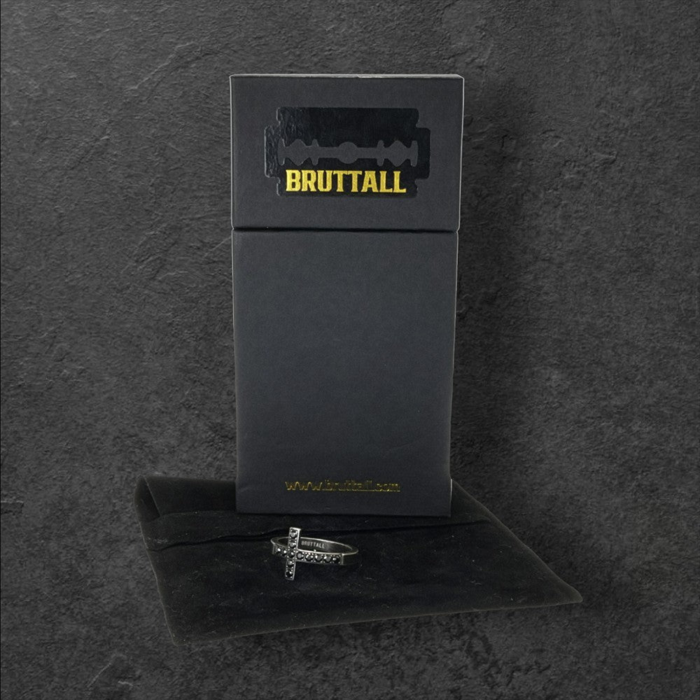 A black box with “BRUTTALL” in bold yellow text and a razor blade design on top displays the TOWER cross Kreuz Ring in antique schwarz, an edgy Schmuckstück resting on a black velvet pouch, all set against a dark background.