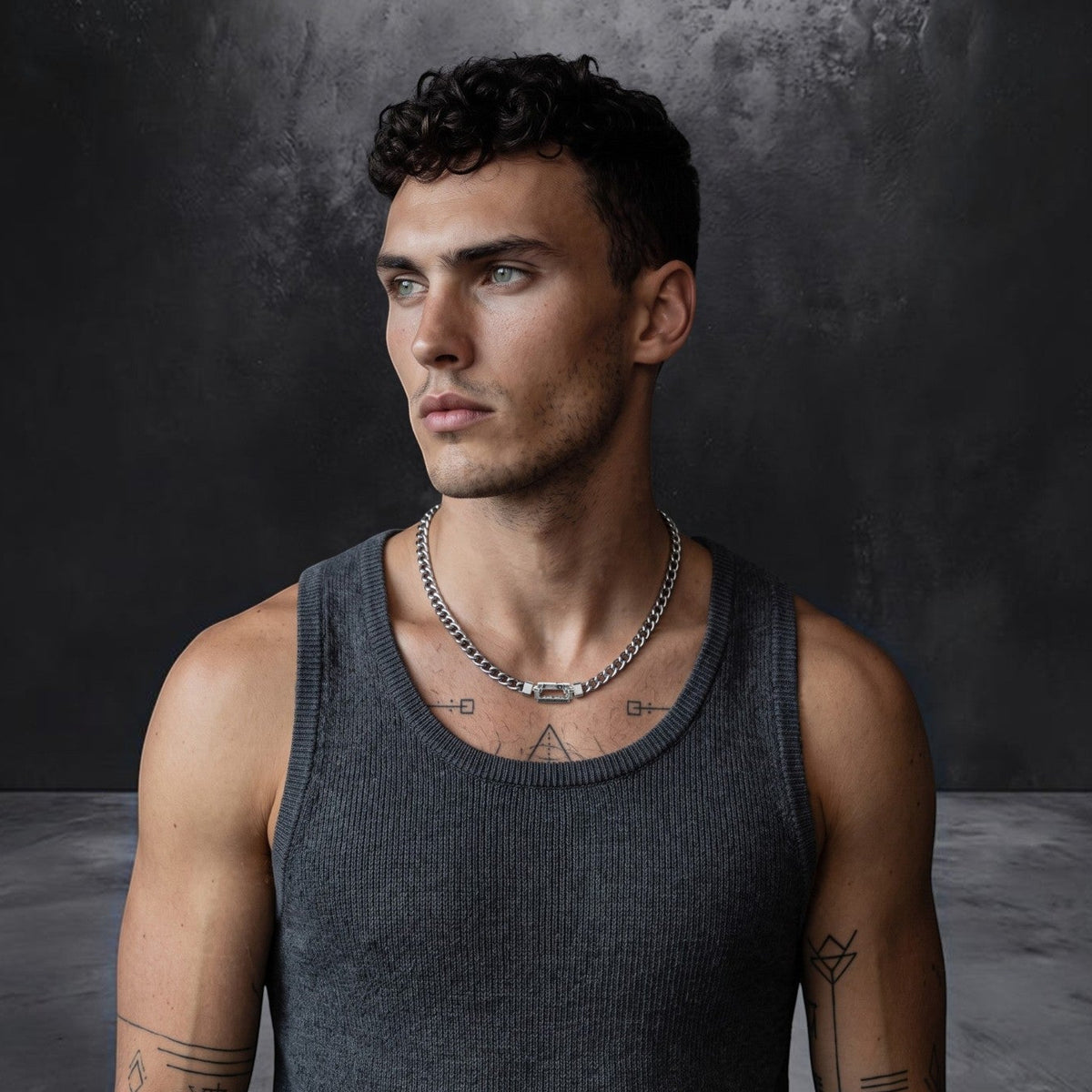 Ein Mann mit lockigem Haar und Tattoos, der ein graues Tanktop und eine Kette von Bruttall Herrenschmuck trägt, die mit Swarovski-Zirkonia verziert ist, steht vor einem strukturierten dunklen Hintergrund und blickt zur Seite.
