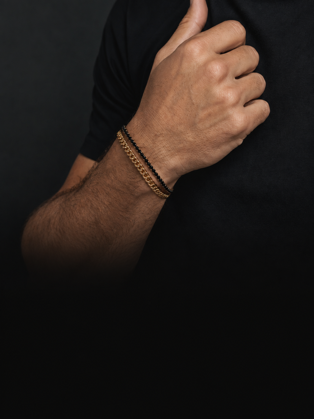Eine Person in einem schwarzen Hemd streckt den Daumen nach oben und trägt zwei Armbänder am Handgelenk: eine goldene Kette und ein schwarzes Perlenarmband, die beide den unverwechselbaren Stil von Bruttall Herrenschmuck widerspiegeln und mit funkelnden Swarovski-Kristallen verziert sind.